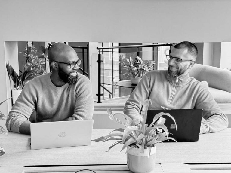 Two employees sit at a desk with their laptops.