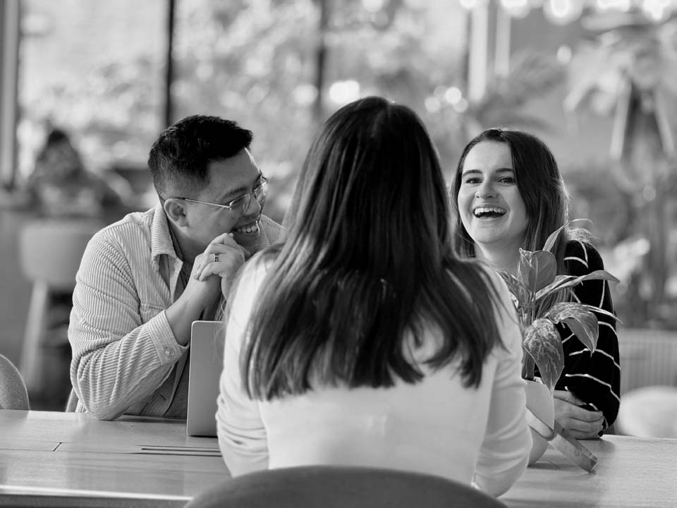 3 colleagues chat around a table and laugh together.