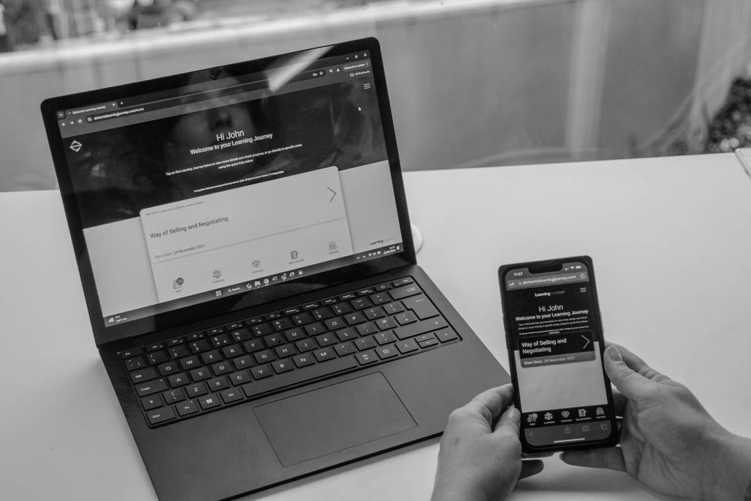 A photograph of a laptop on a desk and a mobile phone in someone's hand - both are showing the Alchemist Learning Journey app.