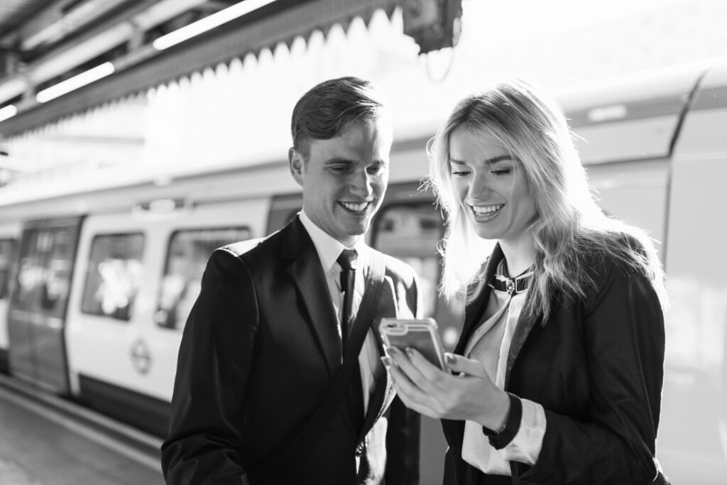 Two people on a train platform smile while they look at a phone.